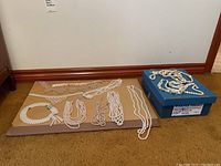 Wide shot of multiple faux pearl necklaces laid out on cardboard and some in a blue box on carpet floor