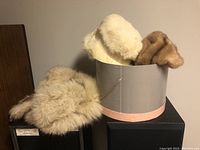 Two fur hats with fur collar and fur wrist pieces placed on and beside black boxes, one hat cream color with satin bow, the other brown mink fur partially in box.