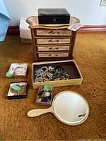 Photo showing large engraved wooden jewelry box, smaller black box on top, open box containing assorted broken costume jewelry pieces, small boxes with earrings, and handheld mirror on floor.