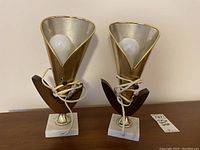 Pair of vintage mid-century modern bedside table lamps with gold metallic cone shades, wooden bases, and white square platforms. Both have electrical cords wrapped at base and bulbs installed.