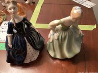 Two Royal Doulton figurines on a wooden table: Debbie HN 2385 in a dark blue dress and Fair Maiden HN 2211 in a pale green dress, viewed from the front and side.