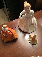 Photo showing three Royal Doulton figurines of ladies in period dresses from above on wooden table.