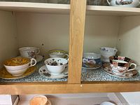 Seven vintage teacups and saucers arranged on two shelves with varied floral and ornate designs.