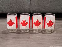 Front view of 4 clear glasses, each featuring a red and white Canadian flag with a maple leaf symbol wrapped around the outside surface.