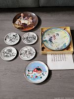 Photo showing all seven plates arranged on a floor including four black and white Norman Rockwell style plates, two Christmas plates, and a colorful horse scene plate in a box.