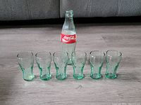 One shot showing one Coca-Cola bottle and six small green-tinted Coca-Cola glasses arranged in a row on a floor, showing bottle label and glass shapes.