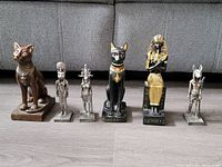 Grouping of six Egyptian-themed statues on floor in front of couch in natural light. The statues vary in material and height between 6 and 9 inches.