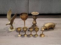 Photo shows all six brass items together on a surface: eagle figurine, pig figurine, large goblet, ornate pricket candle holder, and four stemware glasses.