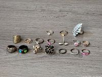 Photo showing all 17 vintage costume rings arranged on a wooden surface. Rings include various metal tones, faux stones in clear, pink, purple, and black cluster styles, two black rings with colorful hearts, and a silver-tone leaf-shaped ring.