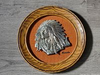 Top-down view of the Native themed wall hanging showing the silver metal chief head relief mounted on orange textured fabric inside circular wood grain frame.