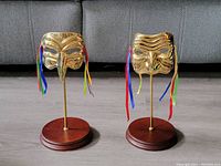Front view of two brass masks on wooden stands showing facial features and colorful ribbons