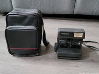 View of the Polaroid OneStep Closeup camera beside its black carrying case on the floor. The camera has an attached strap and the carrying case has two compartments.