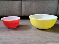Two vintage Pyrex mixing bowls shown from side angle on wood floor with beige couch background. Large yellow bowl on right, smaller red bowl on left.