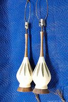 Pair of mid century table lamps shown full view against blue padding, highlighting ceramic base, wooden neck, and metal bulb holder.