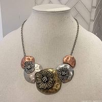 Front view of vintage mixed metal necklace on a display bust, showing three metal plates with floral designs in silver, brass, and copper tones connected by a silver chain.