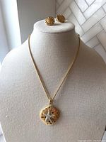 Photo showing the necklace displayed on a bust with the matching clip-on earrings positioned above it, highlighting the pendant design and the sunburst pattern on earrings.