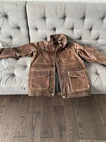 Front view of brown vintage leather jacket laid flat on couch, showing two large front pockets, standing collar, and front zipper hidden behind a flap with snap buttons.