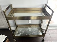 Full view of the brass bar cart showing the rectangular frame with brass finish and mirrored shelves, positioned against wall.