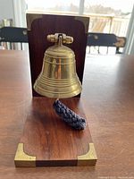 Front angled view of a polished brass bell mounted on a wooden L-shaped base with brass corner protectors. The bell has horizontal ridges and a braided pull rope.