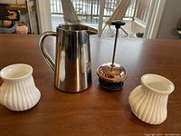 Stainless steel coffee press with copper top plunger and two white Alessi espresso cups on wooden table.