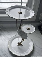 Front angle view of the crane tea server on a marble surface, highlighting the three porcelain plates attached to the crane figure with metal legs and neck.