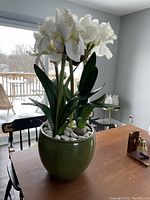 Faux white flower plant in green planter on wooden table with snowy outdoor background visible through window.