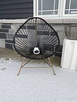 Black vinyl Acapulco chair on rose gold metal frame against stone wall on snow-covered ground.