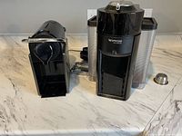 Photo showing both Nespresso machines placed side by side on a marble countertop, highlighting their size difference and exterior condition.