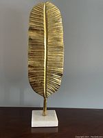 Front view of gold metal palm leaf sculpture with black stem on white marble base