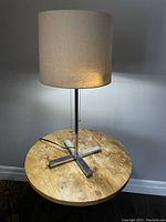 Front angle of the lamp showing chrome X-shaped base, beige Belgian linen drum shade, and cord with pull chain switch.