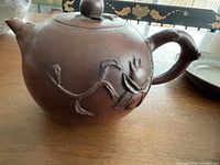 Side view of dark brown teapot showing rounded body and floral relief design with curving stems and flower.