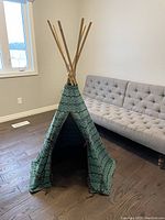 Full view of the children's tepee showing the entrance and fabric pattern in teal and black set up in a room next to a sofa.