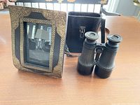 Overall view of binoculars, case, and carved brass photo frame on wooden table