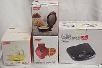 Four boxed items stacked: Dash Rapid Egg Cooker (yellow), Dash Mini Maker Griddle (red), Dash Heart Mini Waffle Maker (red, heart shaped), and Flat Grill Sandwich Maker (black/silver)