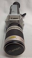 Front three-quarter view showing Canon badge on top plate and Auto Zoom 1014 front label, plus front of zoom lens with Canon lens cap and Passed quality sticker.