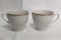 Pair of white porcelain tea cups with gold rim and handles shown from side angle.