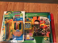 Kids coloring and painting supplies lot laid out on wooden floor showing paintbrushes, colored pencils, paint palette, coloring books, and playing cards.