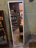 Tall rectangular white-framed full-length mirror leaning against concrete wall