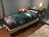 Side angle of twin bed showing thick mattress and Mario themed fitted sheet with plush toys