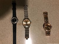 Three women's wristwatches laid out on a beige surface: Worthington with black rubber band, silver-tone watch with metal band, rose gold-tone Merona watch with rhinestones.