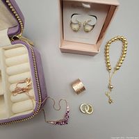 Top down photo showing all items: rose gold necklace with purple gemstones, gold-tone cubic zirconia earrings in pink box, textured silver ring, adjustable silver bracelet, velvet jewellery box