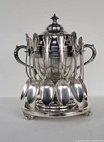 Front view of silver plated spooner with 12 spoons hanging around top edge, showing detailed handles and body shape