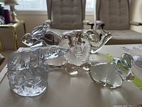 Photo shows six clear glass animal figurines arranged on a flat surface with some reflecting light, sitting in front of a window with chairs in background.