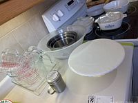 Overall lot display of measuring cups, metal shaker, mixing bowls, cake stand, and casserole dish on kitchen countertop.