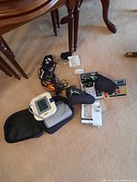 Lot photo showing Omron blood pressure monitor, calculators, small speaker, shaver, headphones, and cords on carpeted floor.