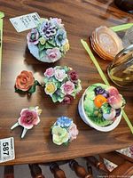 Photo showing multiple bone china floral figurines in bright colors arranged on a table including roses and pansies alongside a fruit-themed trinket jar.