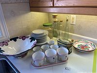 Overview of white china cups, plates, saucers, large painted bowl, glass bottles with corks, and metal biscuit tin shown on kitchen counter.