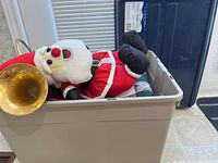 Storage bin partially open showing a large Santa Claus plush in red and white costume, black boots, and gold horn peeking out of the bin.