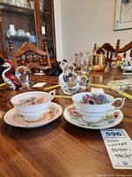 Two teacups with matching saucers showing different floral patterns and gold trim, placed on a wood table alongside other glass items.