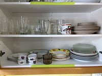 Two shelves view showing assorted clear glassware including water glasses and mugs, various plates and saucers with floral and fruit patterns.